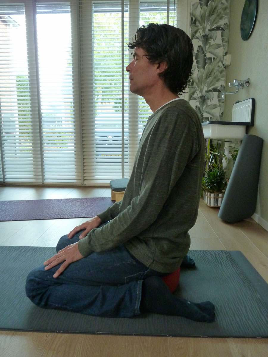 Knielend op een rood meditatiekussen tussen de benen, met de onderbenen naar achteren op de mat
